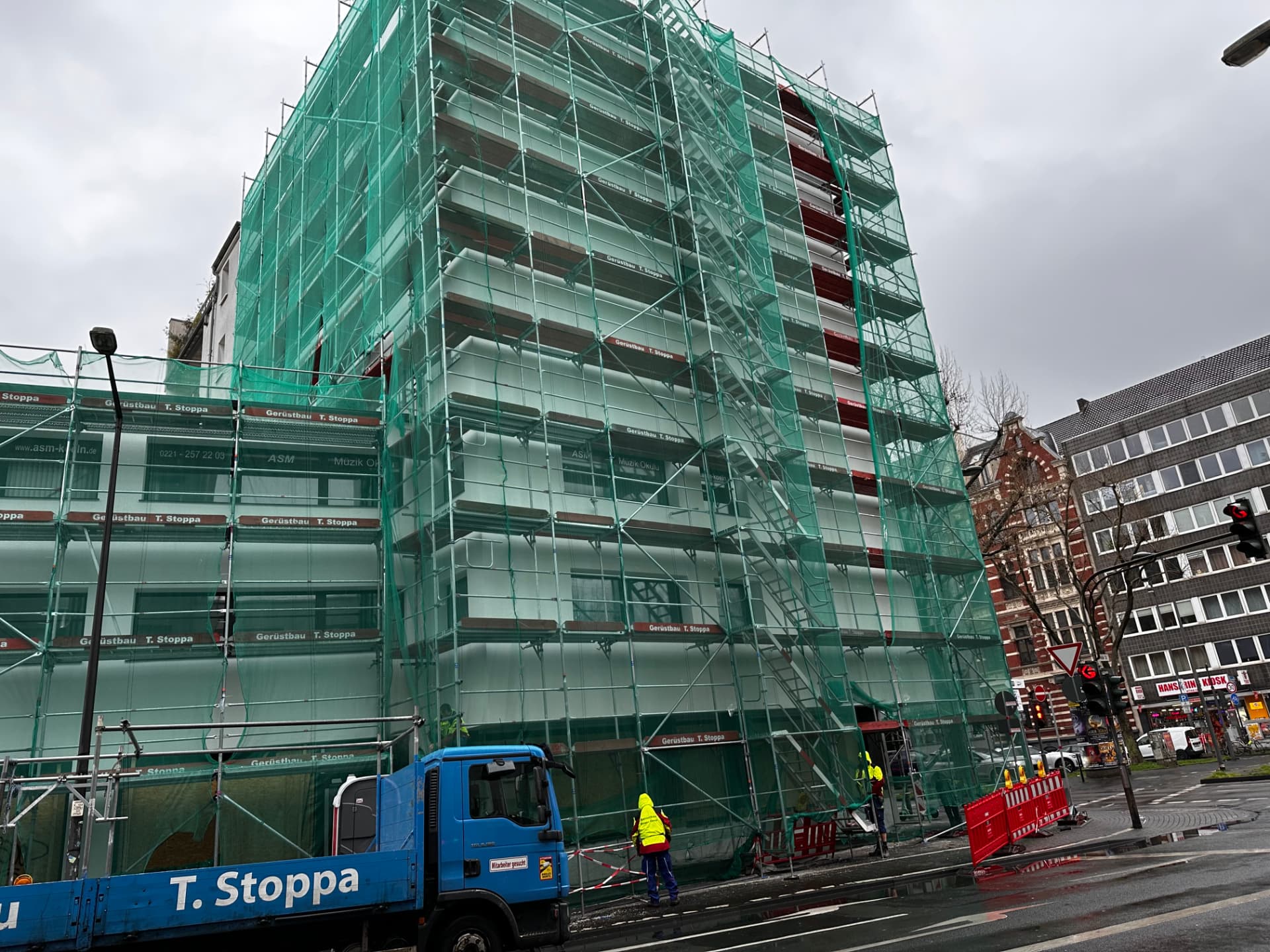 Büro- und Geschäftshaus am Hansaring in Köln vollflächig eingrüstet mit grünen Staubschutznetzen und Treppenturm-Gerüst von Gerüstbau Stoppa, blauer Stoppa-Lkw davor