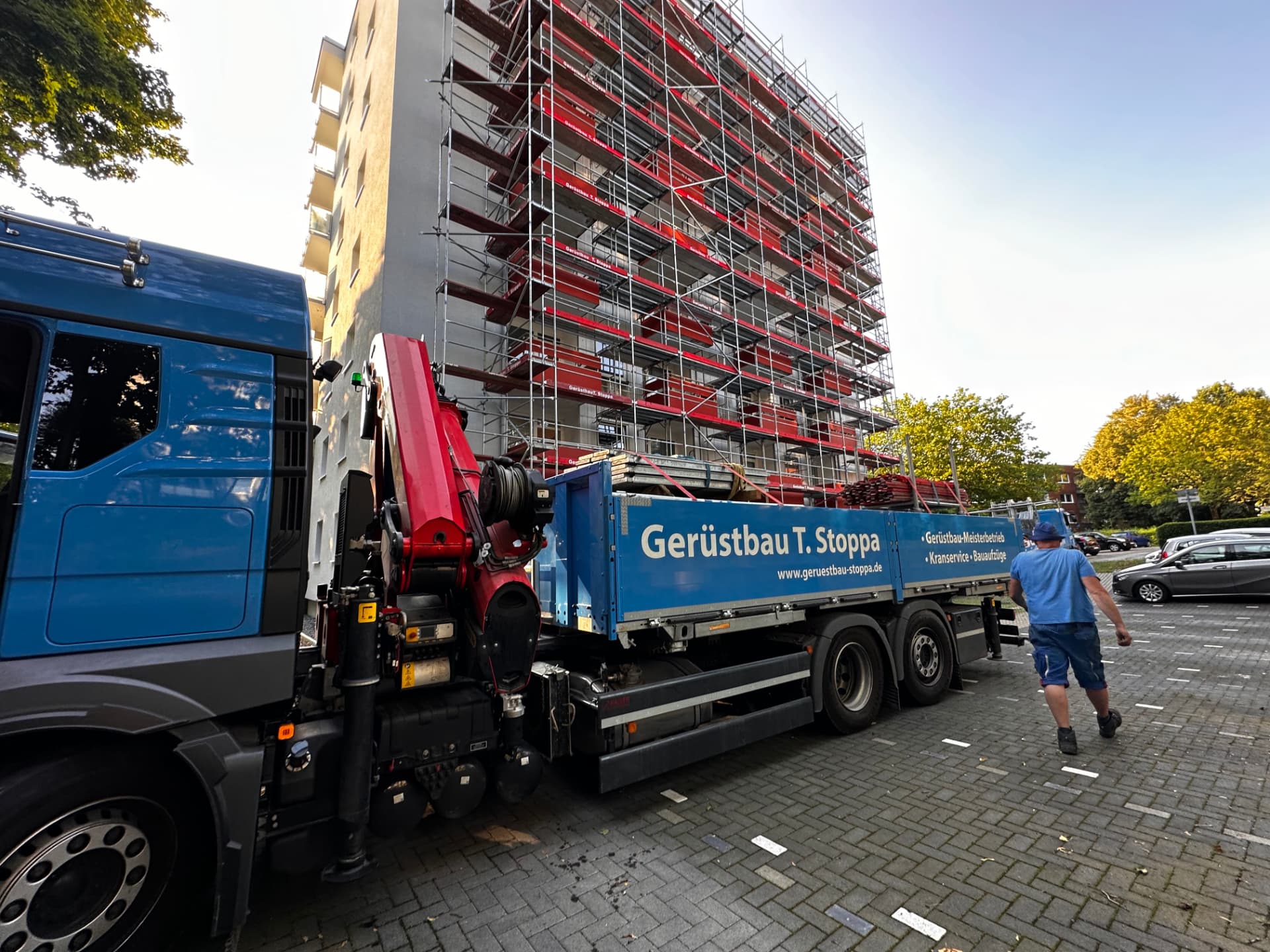 Gerüstbau-Stoppa-Lkw mit rotem Ladekran entlädt Gerüstmaterial vor einem voll eingerüsteten Wohnhochhaus